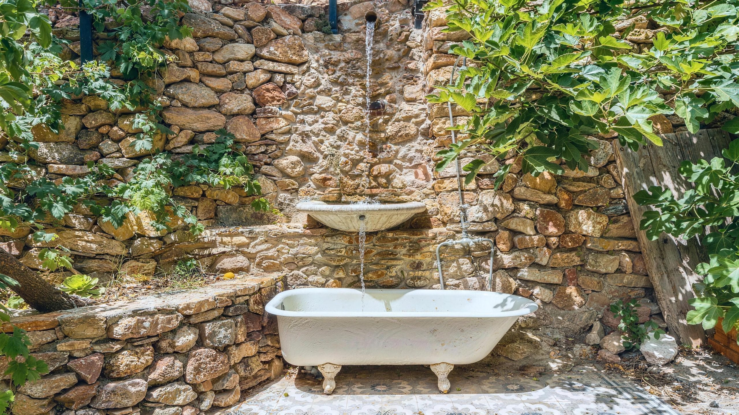 Bañera exenta de porcelana al aire libre, con el caño saliendo del muro de piedra seca y la higuera dando sombra, en el jardín de la Casa Museo del Trovo y Aguardiente de Murtas