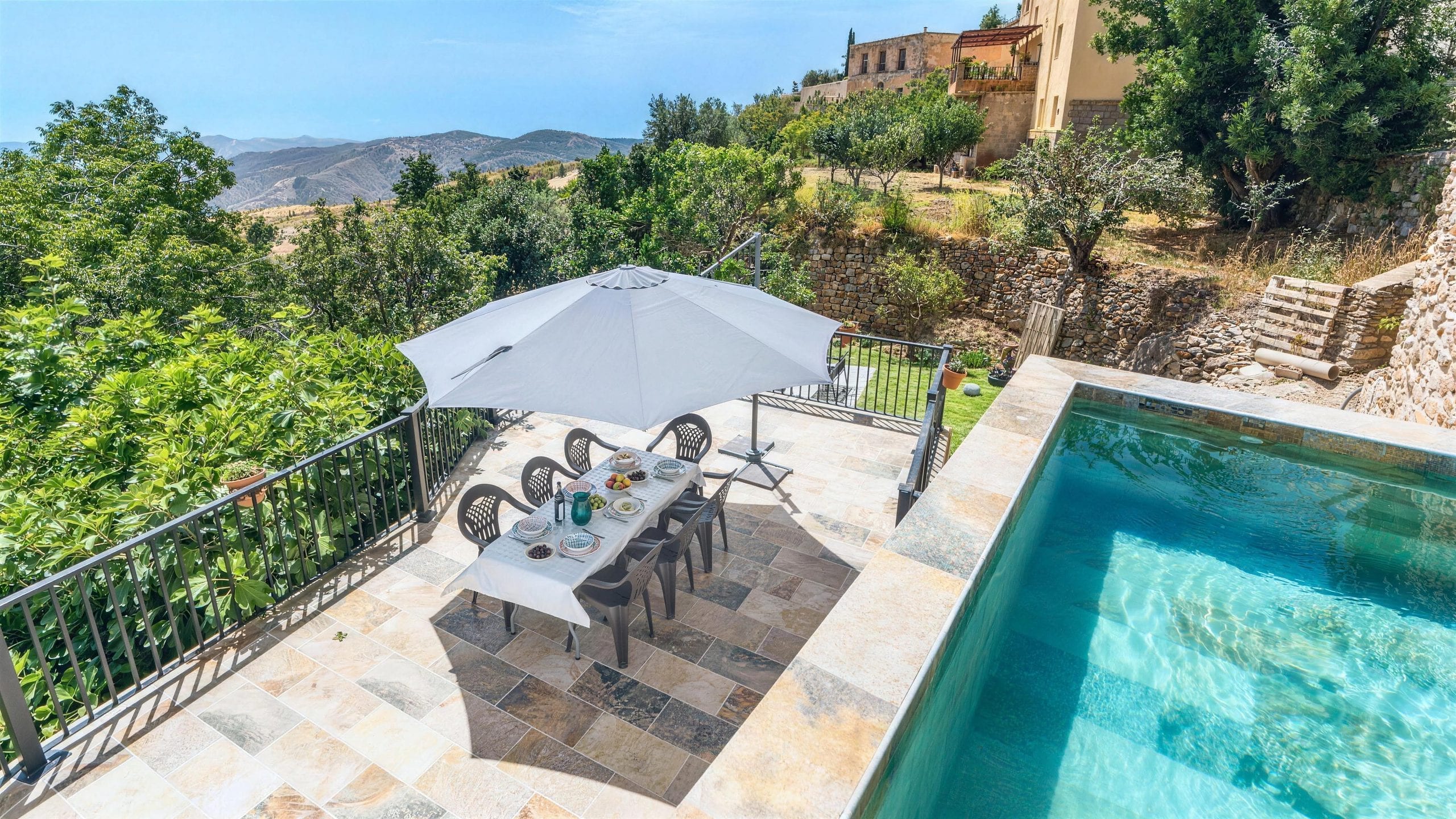 Piscina y mesa larga bajo la sombrilla, con los bancales de olivos y el valle detrás, en la Casa Museo del Trovo y Aguardiente de Murtas